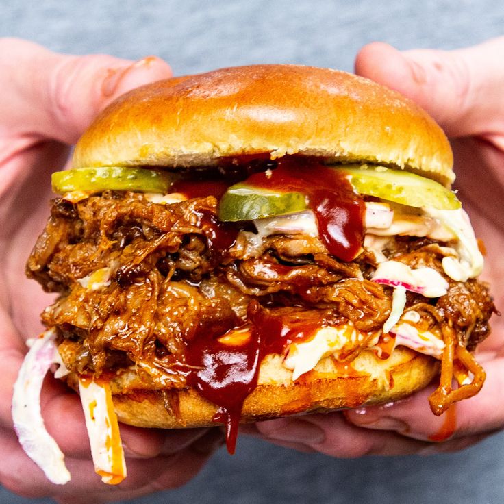 a hand holding a pulled pork sandwich with ketchup and pickles on it