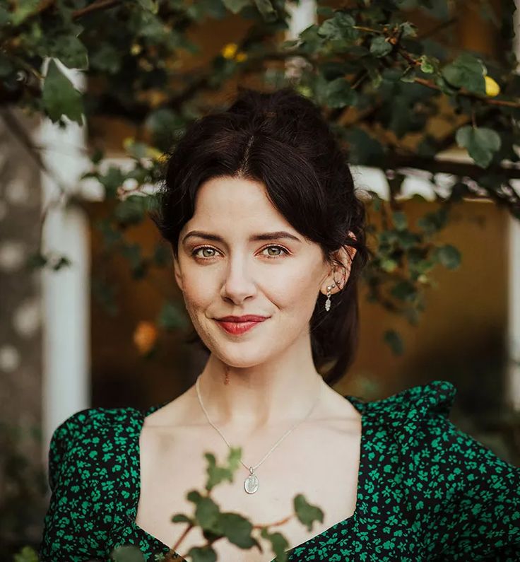 a woman wearing a green dress standing in front of a tree and looking at the camera