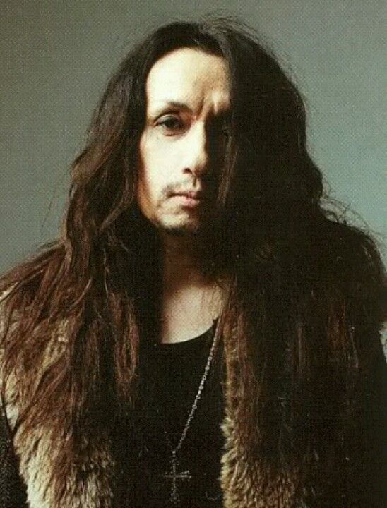 a man with long hair wearing a black shirt and fur vest, looking at the camera