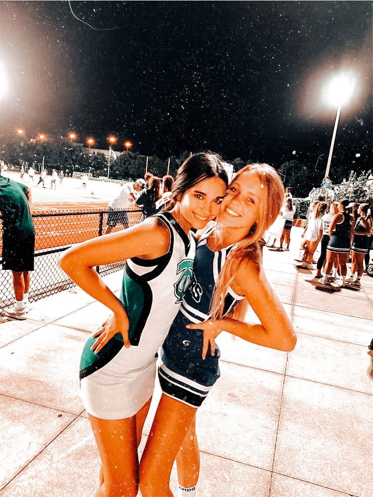 two young women hugging each other in front of a baseball field at night with lights on