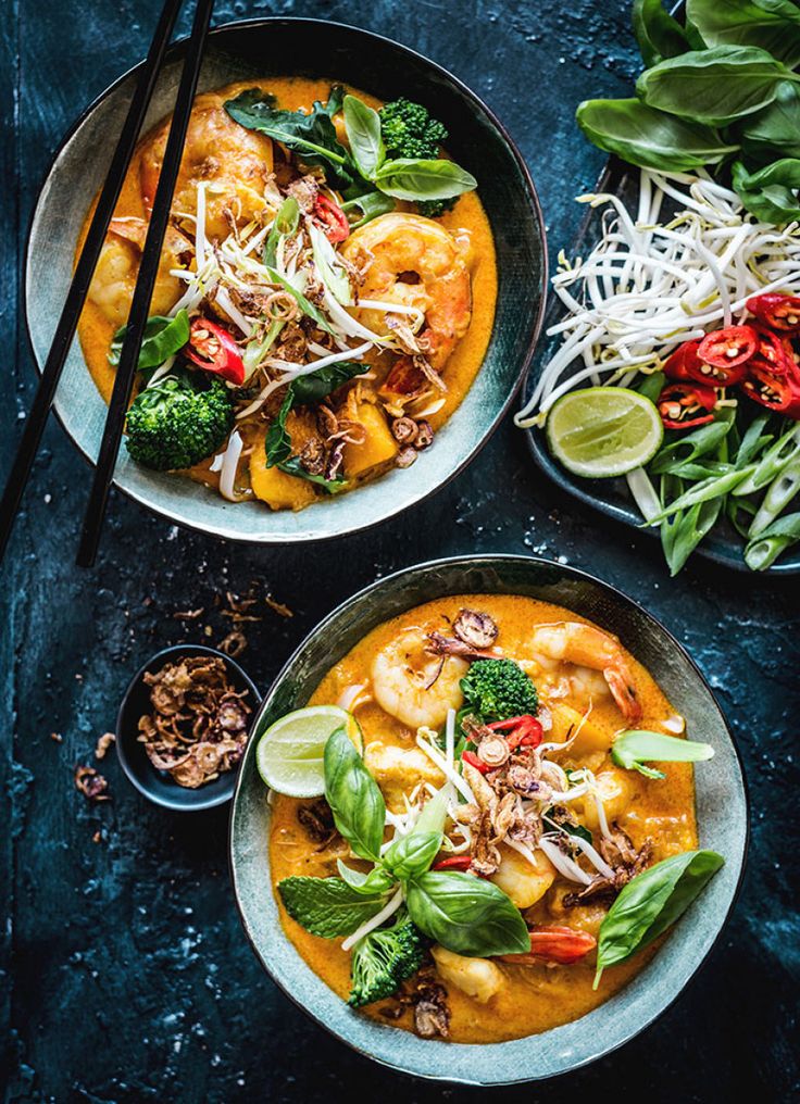 two bowls filled with shrimp and vegetables next to chopsticks on a blue surface