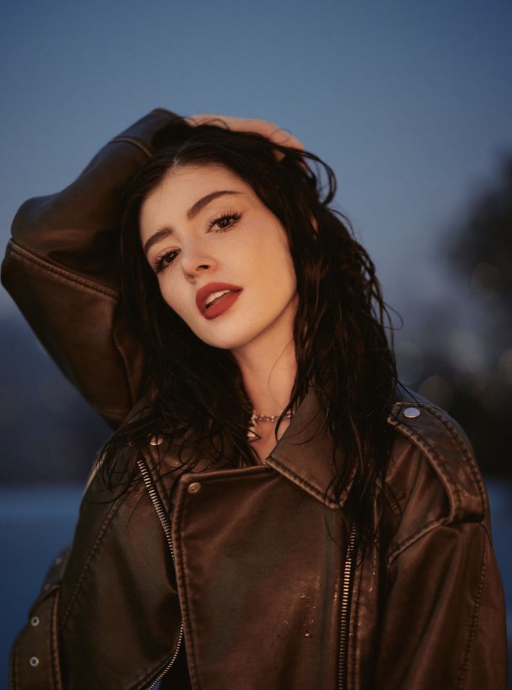 a woman with long hair wearing a leather jacket and posing for the camera at dusk