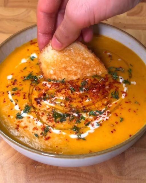 a hand dipping some bread into a bowl of soup with cheese and herbs on top