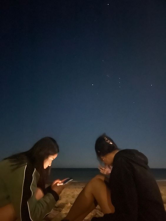 two people sitting on the beach looking at their cell phones