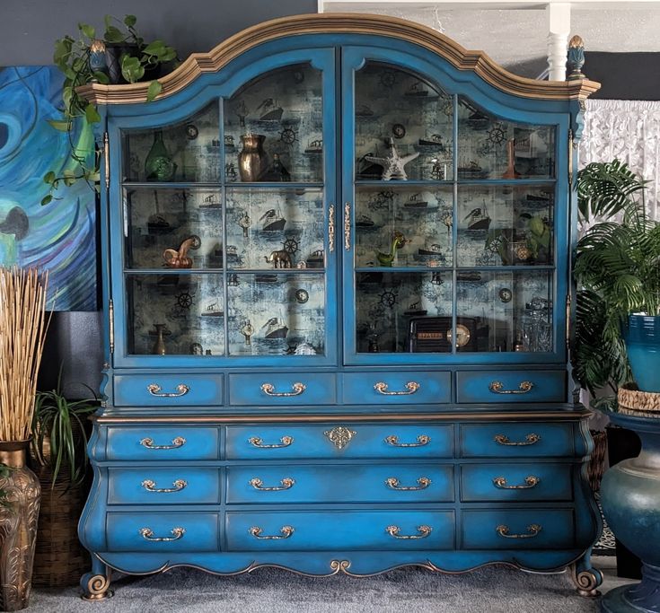 a blue china cabinet with glass doors and drawers in front of a painting on the wall