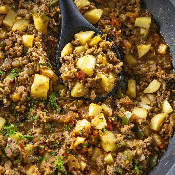 a skillet filled with meat and potatoes on top of a table next to a wooden spoon