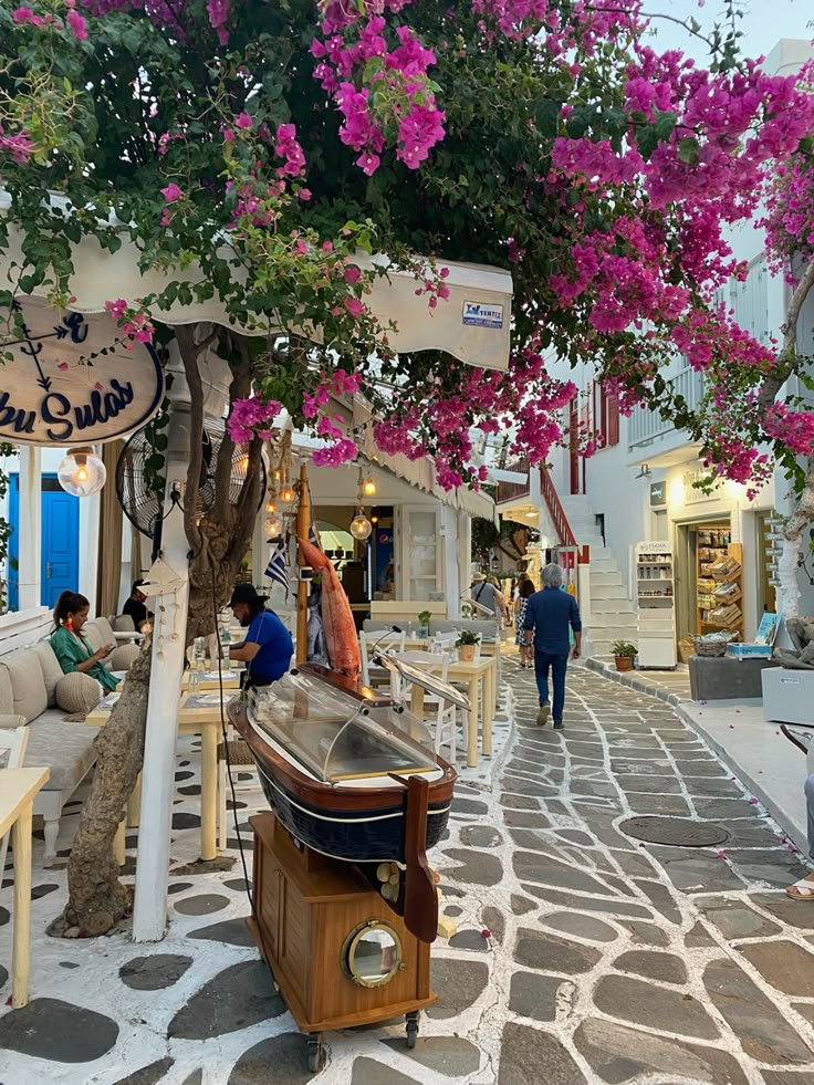 people are walking down the street in front of shops with pink flowers hanging from them