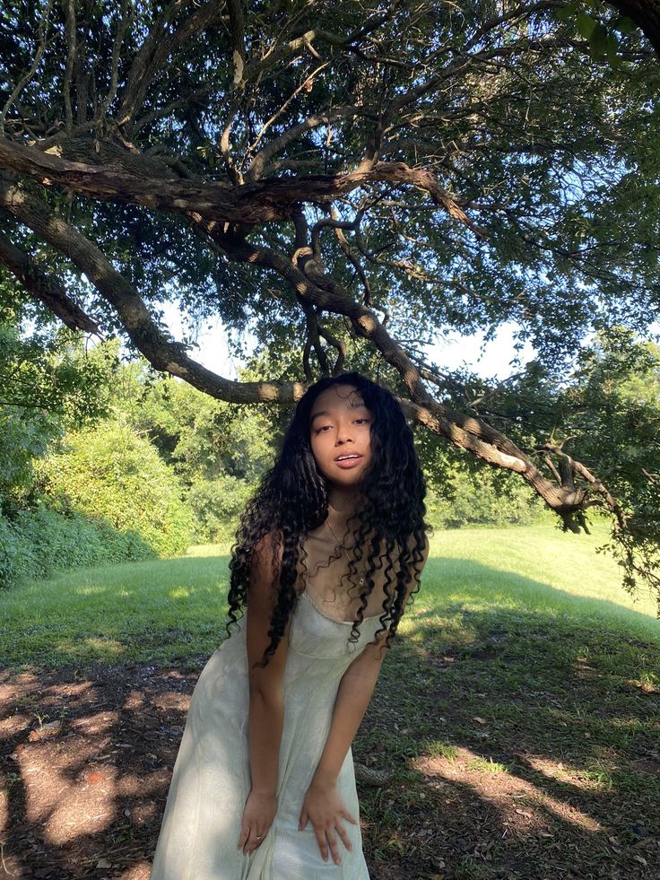 a woman in a white dress standing under a tree with her hands on her hips