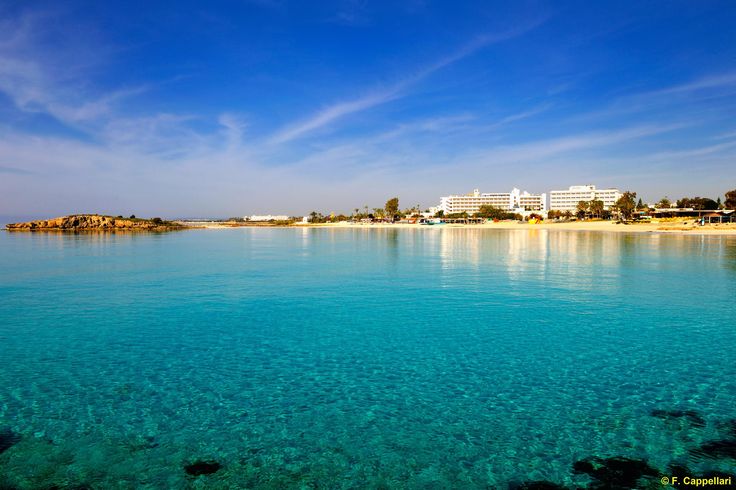 clear blue water with white buildings in the background and sand on the beach behind it