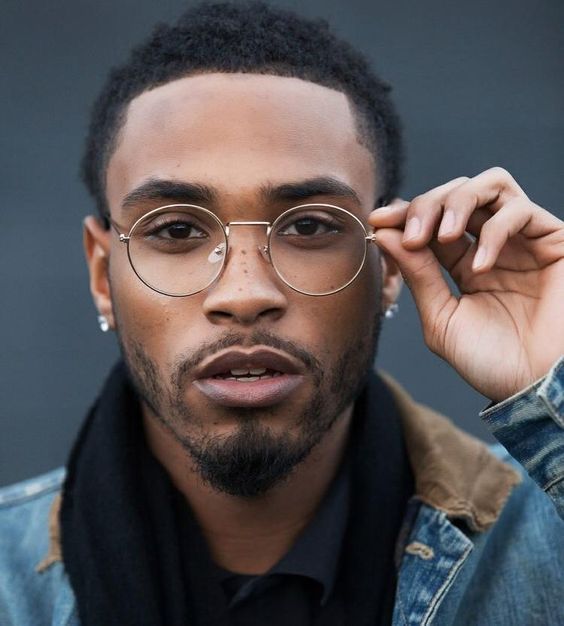 a man wearing glasses and a denim jacket is looking at the camera with his hand on his head