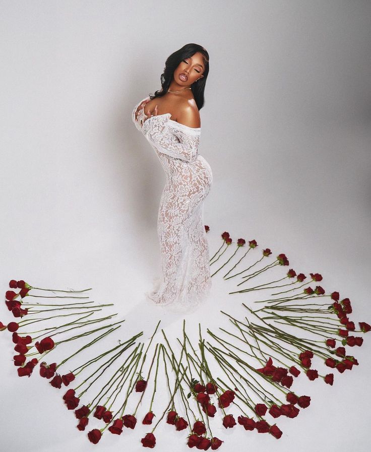 a woman in a white dress standing next to red flowers on a white surface with her arms behind her back