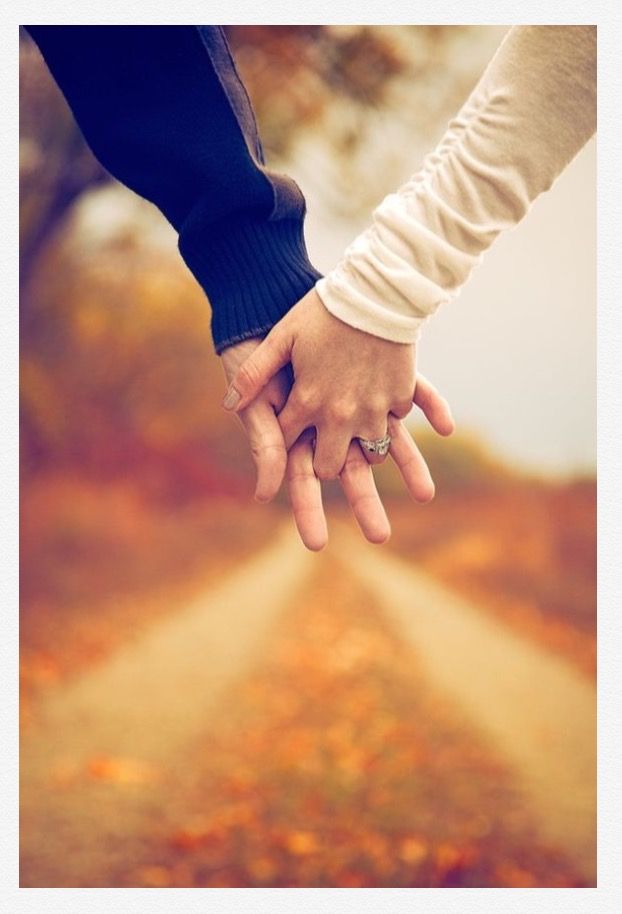 two people holding hands while walking down a dirt road in the fall with leaves on the ground