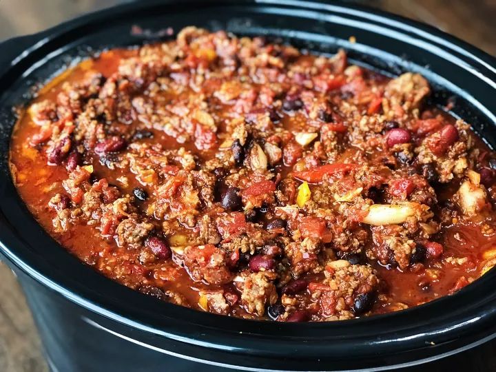 a crock pot filled with chili and meat in it sitting on a wooden table