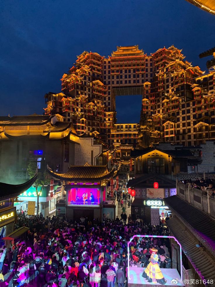 a large group of people standing around in front of buildings at night with lights on them