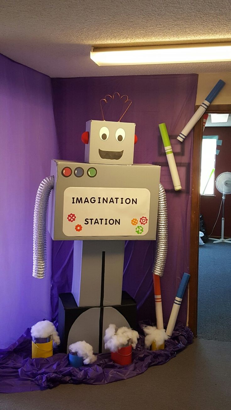 a cardboard robot standing in front of a purple wall with the words imagine station written on it