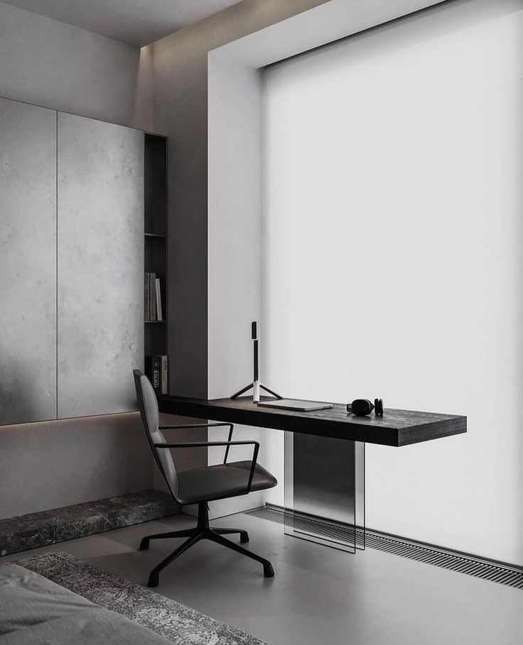 a black and white photo of a desk in a room with large windows, an office chair and a book shelf