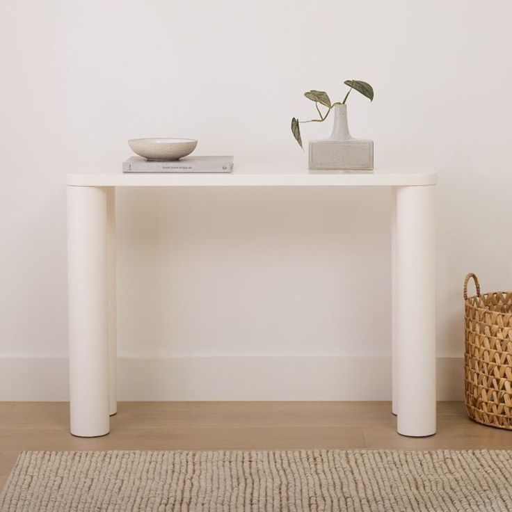 a white table with a plant on top and a basket next to it in the corner