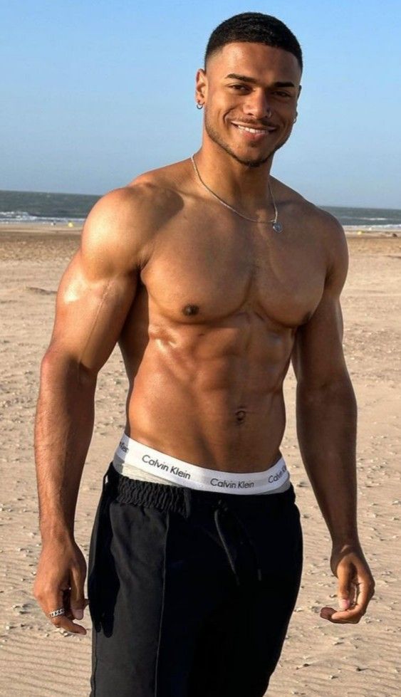 a man standing on top of a sandy beach next to the ocean with his shirt off