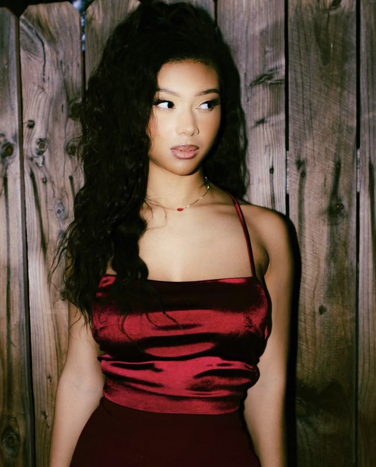 a woman in a red dress standing next to a wooden wall and looking at the camera