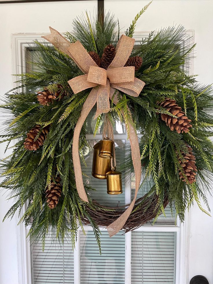 a wreath with two bells hanging from it's side and pine cones on the front door