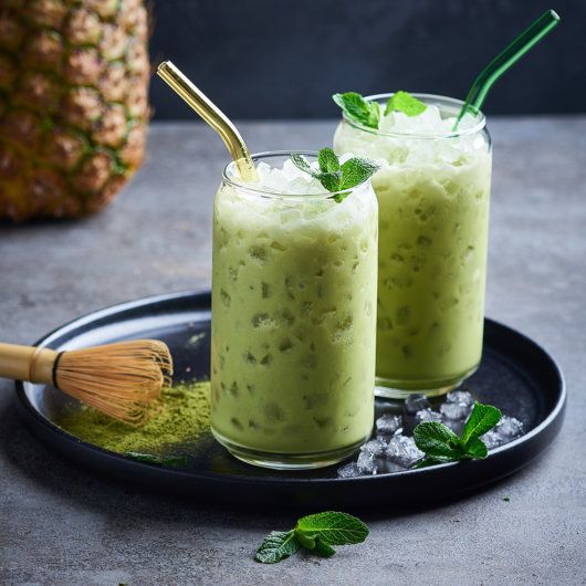 two glasses filled with green smoothie sitting on top of a plate next to a pineapple