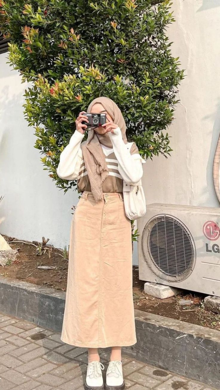 a woman standing on the sidewalk taking a photo with her camera and wearing a hijab