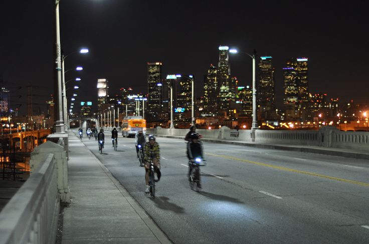 several people riding bikes on a bridge at night with city lights in the back ground