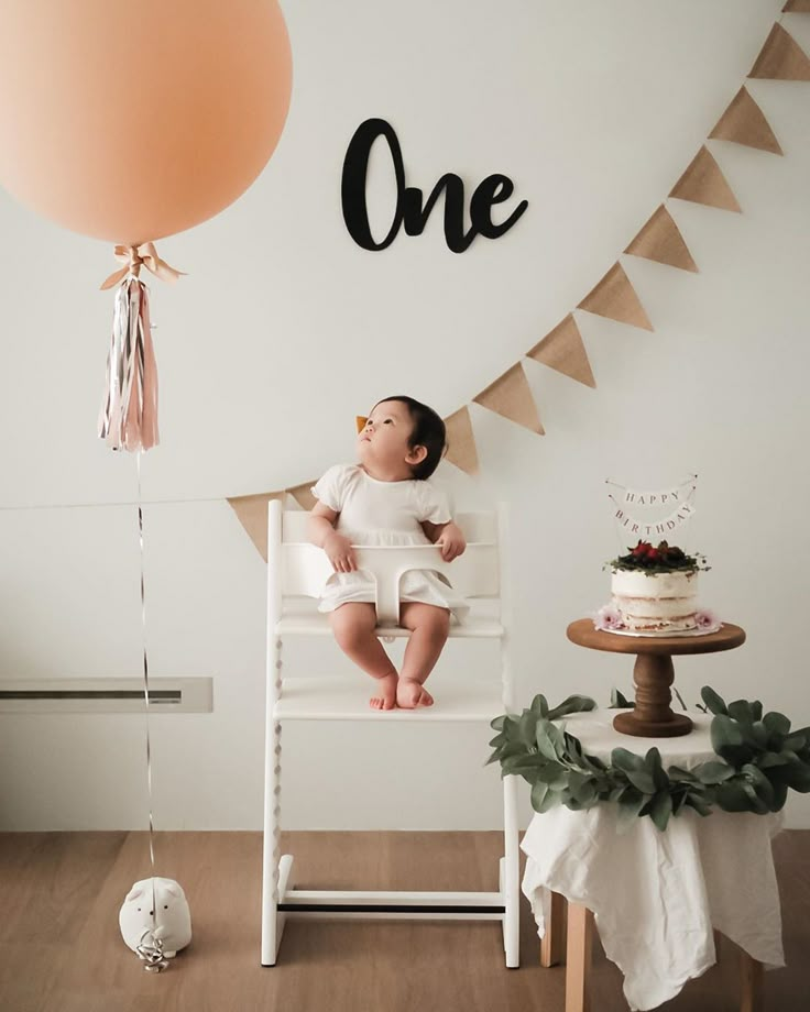 a baby is sitting in a chair with a cake and balloons on the wall behind him