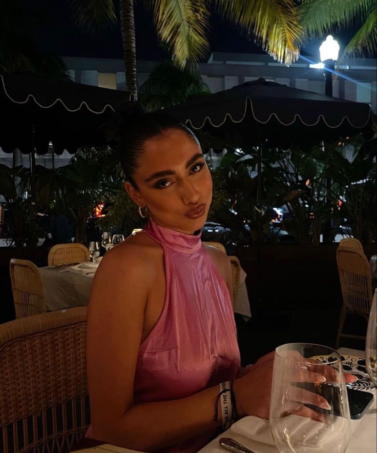 a woman sitting at a table with a glass of wine in front of her and palm trees behind her