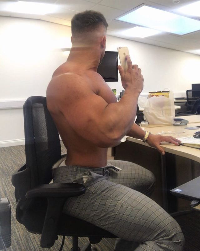 a shirtless man sitting at a desk taking a selfie with his cell phone