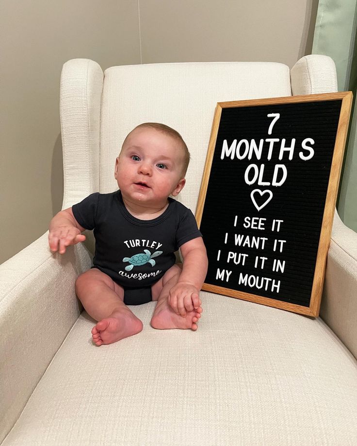 a baby sitting in a chair next to a sign that says, 7 months old i see it i want it but in my mouth