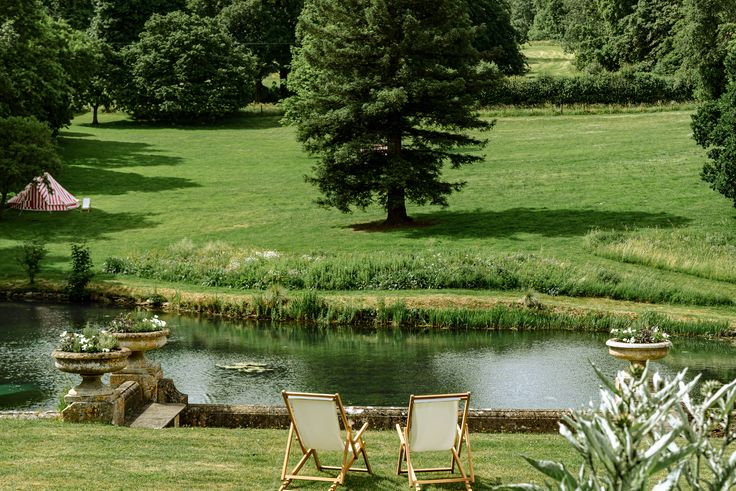 two lawn chairs sitting on top of a lush green field next to a lake and forest