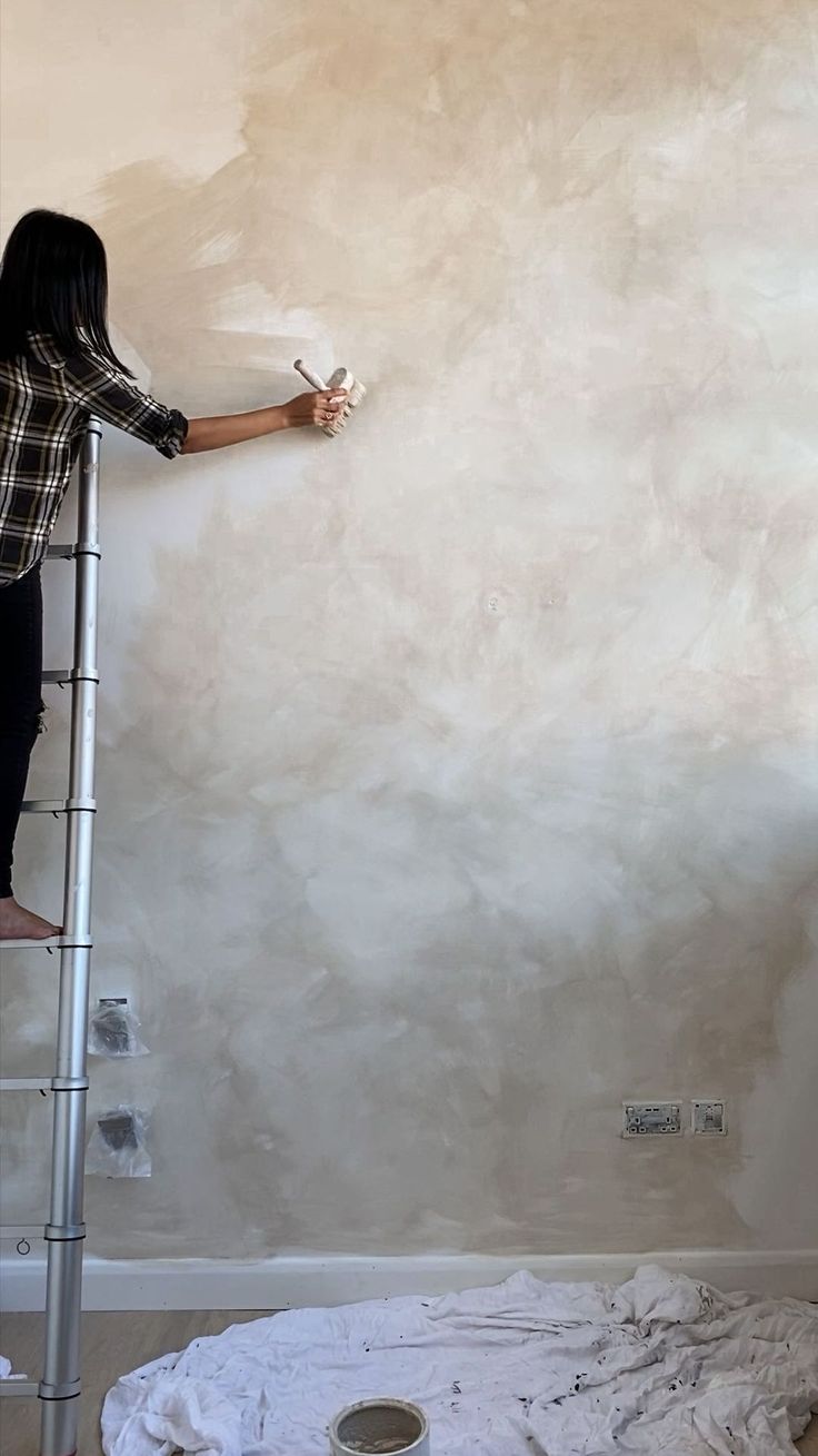 a woman standing on a ladder painting a wall with white paint and holding a brush