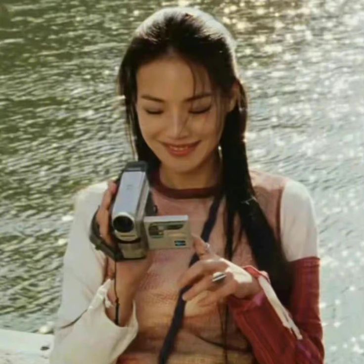 a woman holding a camera in her hand and smiling at it's screen while standing next to the water