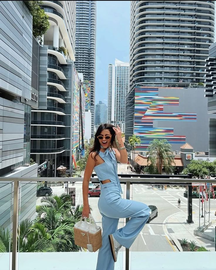 a woman in blue jumpsuits and sunglasses standing on a balcony with buildings behind her