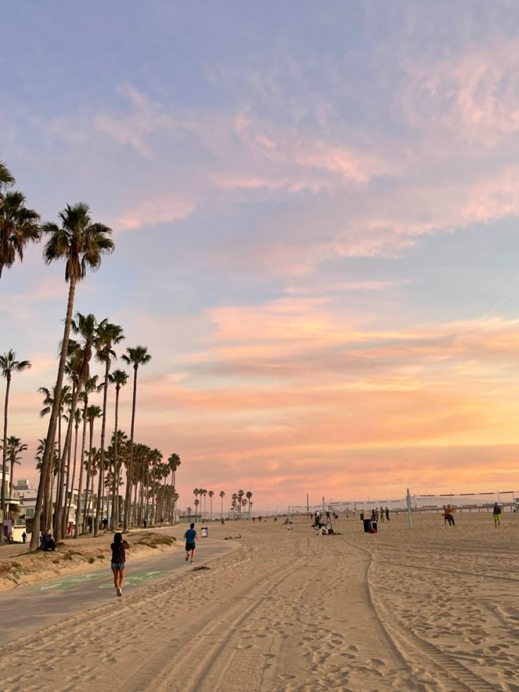 people walking on the beach at sunset with palm trees in the foreground and pink clouds in the sky