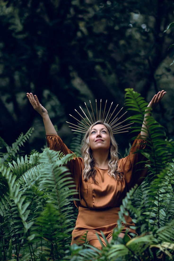 a woman with her hands up in the air surrounded by plants and trees, wearing an orange dress