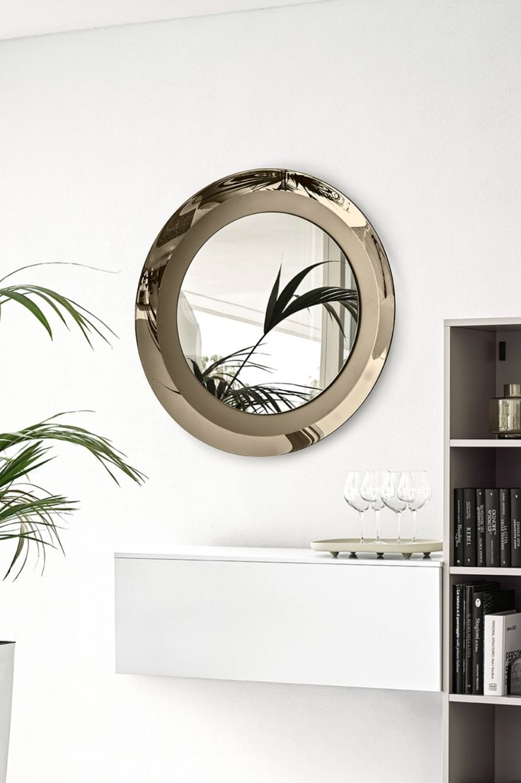 a round mirror hanging on the wall above a white shelf with a potted plant