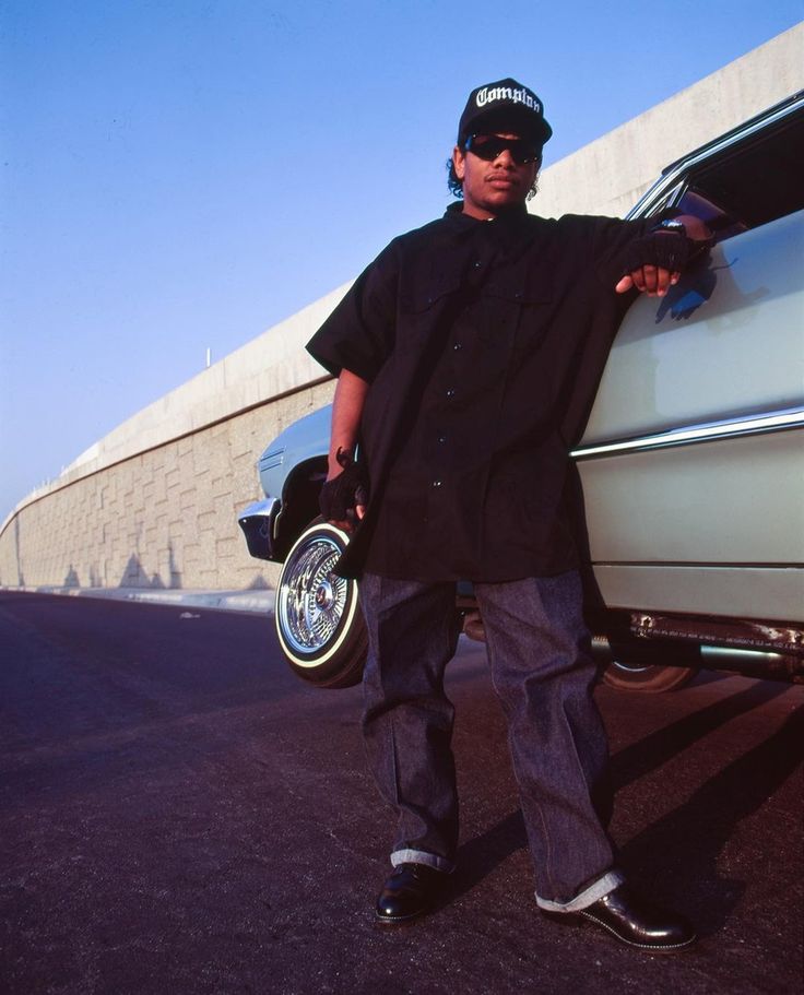 a man standing in front of a car with his hand on the wheel and wearing sunglasses