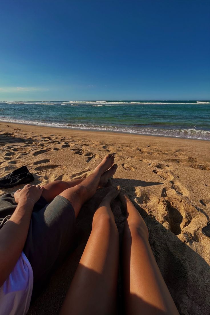 two people are laying on the beach with their feet in the sand looking out at the ocean