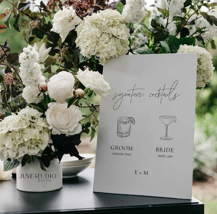 white flowers are in vases next to a sign with the names of bride and groom