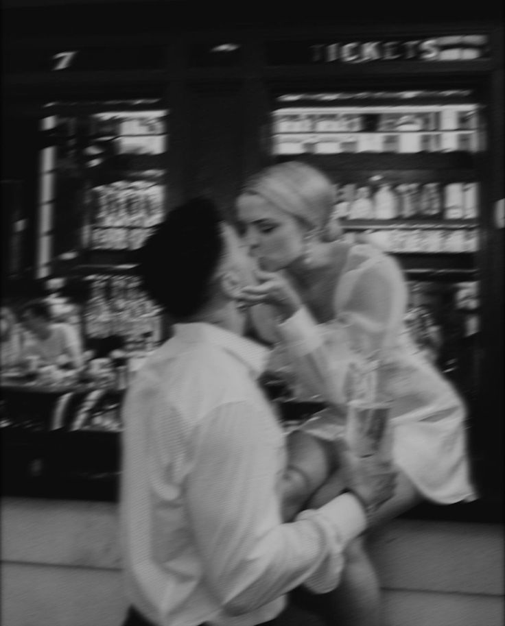 black and white photograph of two people walking in front of storefronts with drinks