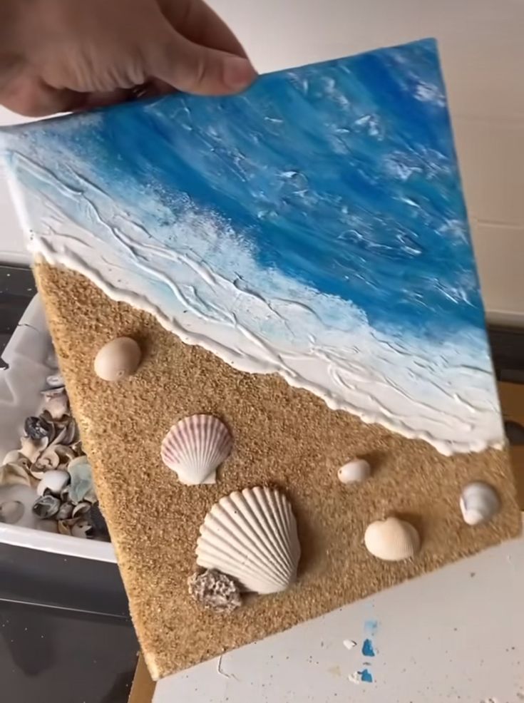 someone is holding up a piece of art with seashells and sand on it