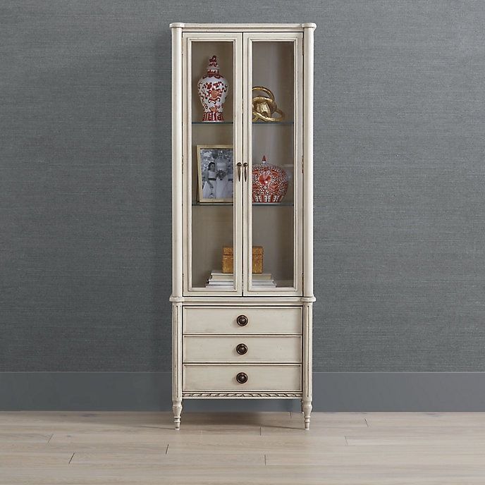 a tall white cabinet with glass doors and drawers on the bottom shelf, in front of a gray wall