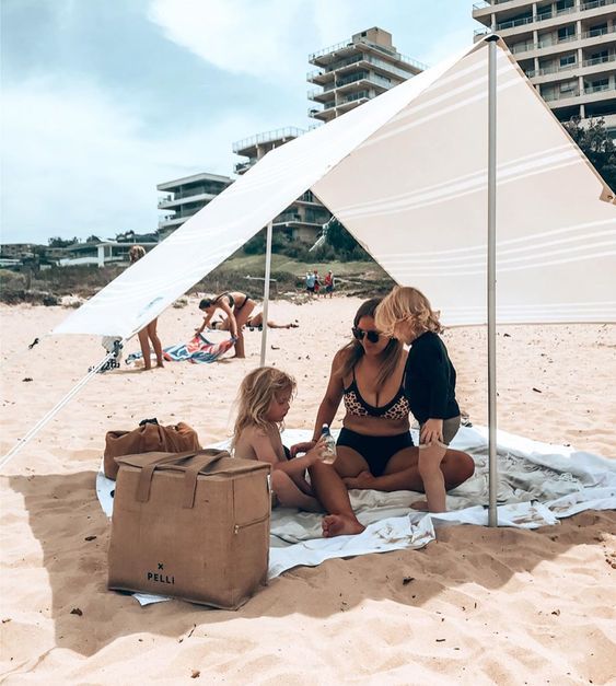 two women and a child sitting on the beach under a white tent with people in the background