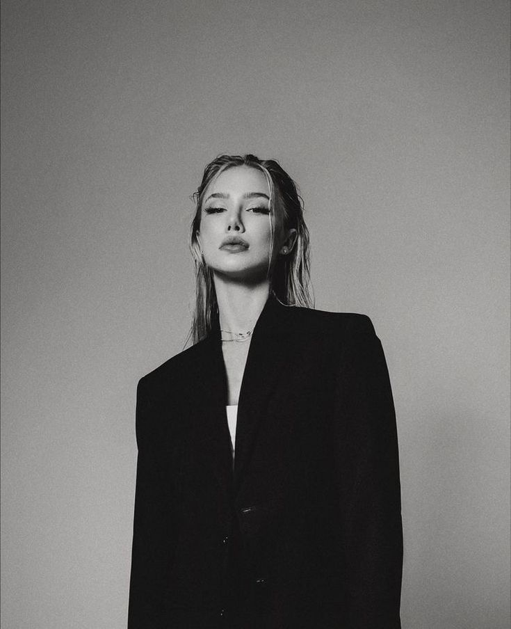 a black and white photo of a woman with long hair wearing a suit, standing in front of a wall