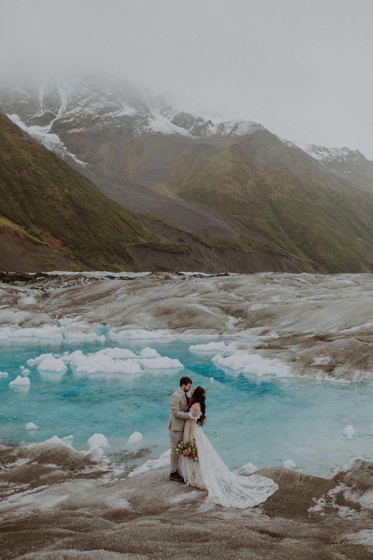 a bride and groom standing in front of a mountain lake with snow on the mountains behind them