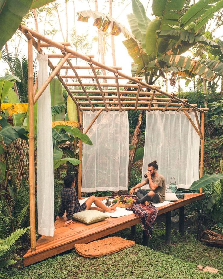 two people sitting on a wooden bench in the middle of a jungle area with white curtains