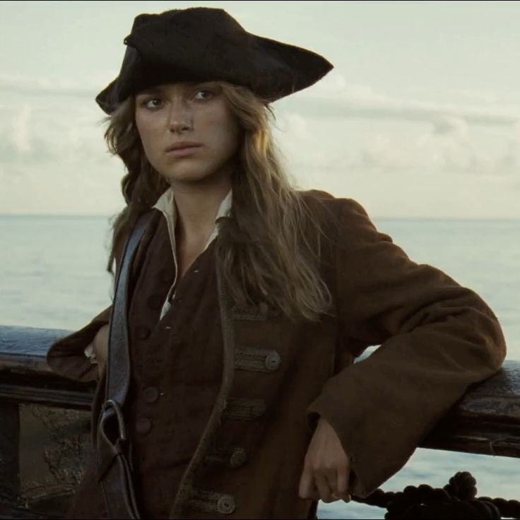 a woman with long hair wearing a black hat and brown jacket standing next to the ocean