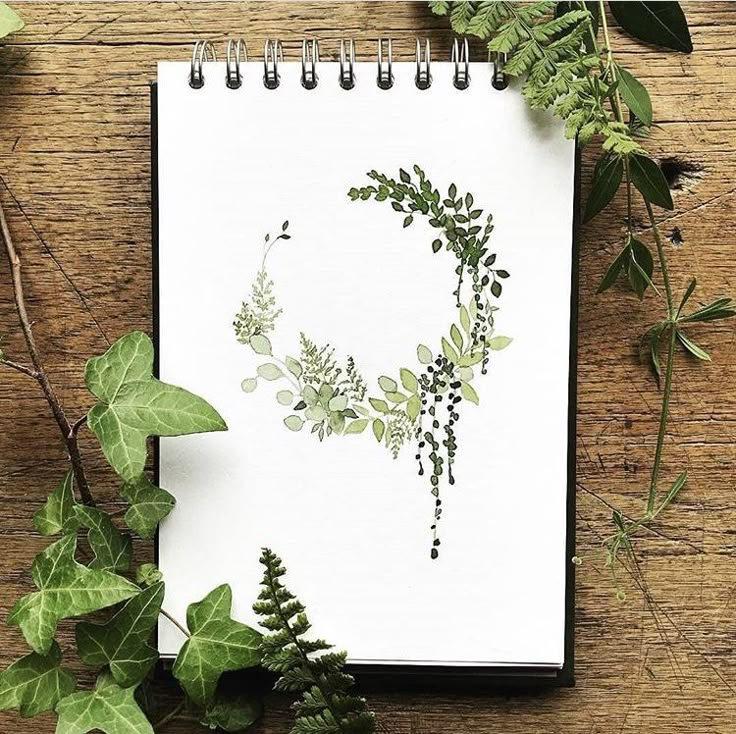 a spiral notebook with green leaves and plants around it on a wooden table next to a plant pot